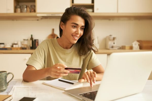 Cute Arabic girl transferring money with online banking using wireless internet connection on laptop. Stylish young woman holding debit card entering personal payment data, purchasing goods distantly